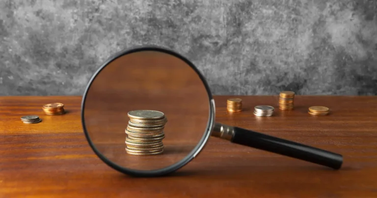 Stack of coins magnified by a lens on a wooden table, symbolizing cost analysis and savings.